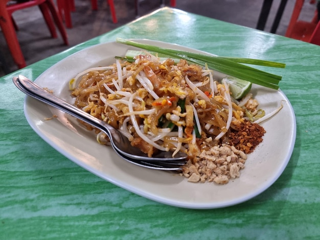 „Pad Thai” Podawany w naczyniu w kolorze skorupki jajka z zieloną cebulą Plastry cytryny Zmielone prażone orzeszki ziemne Łyżka i widelec na zielonym stole