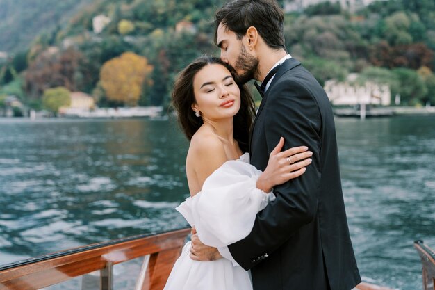 Pan młody przytula pannę młodą i całuje jej głowę stojąc na jachcie pływającym po jeziorze Como we Włoszech