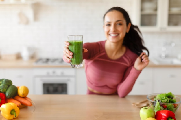 Pani fitness stoi w kuchni i oferuje świeżo przygotowany zielony koktajl