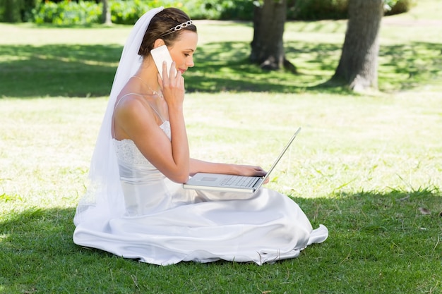 Panna młoda używa laptop i telefon komórkowego w parku