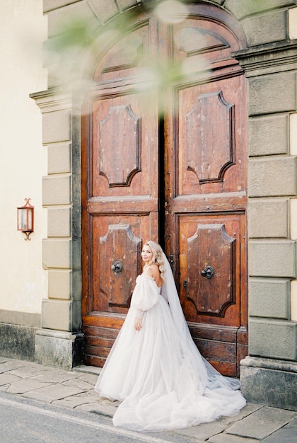 Panna młoda w białej sukni z welonem stoi przy drewnianych drzwiach budynku