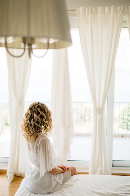 Zdjęcie panna młoda w delikatnym peniuarze siedzi na łóżku przy szerokim oknie tarasowym z białymi zasłonami