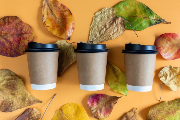 Papierowe kubki do kawy z pokrywkami w jesiennych liściach z pomarańczowym tłem