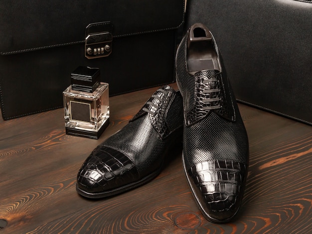 Para czarnych męskich butów leży na powierzchni ciemnych, wyrzuconych desek, obok butelki perfum na tle dwóch skórzanych męskich teczek. Widok z boku. Styl szefa