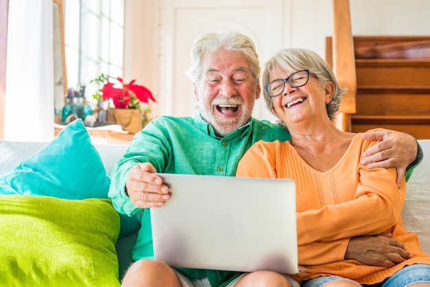 Para dwojga szczęśliwych dojrzałych i starych ludzi lub seniorów w domu siedzących na kanapie, cieszących się i bawiących się razem, patrząc i korzystając z laptopa lub komputera PC