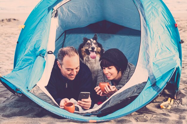 para patrząc na smartfona i bawić się w namiocie na bezpłatnym kempingu na plaży Pies border collie za nimi patrząc w kamerę. vintage kolory i koncepcja rodziny wakacje. alternatywa t