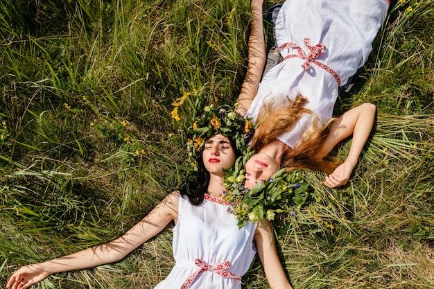 Para pięknych młodych dziewczyn przyjaciół kłama na trawie w lecie i cieszy się życie. Portret dwóch kochających kobiet w słowiańskich tradycyjnych strojach. Przyjaciele spędzają czas w naturze.