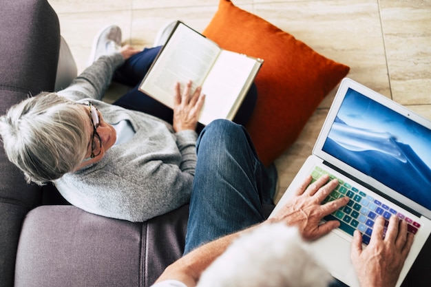 Para Seniorów Razem W Domu - Dojrzały Mężczyzna Pracujący Siedzący Na Kanapie Lub Oglądający Filmy Z żoną Siedzącą Na Ziemi Czytającą Książkę W Ciszy - Kaukaski Na Emeryturze Kryty