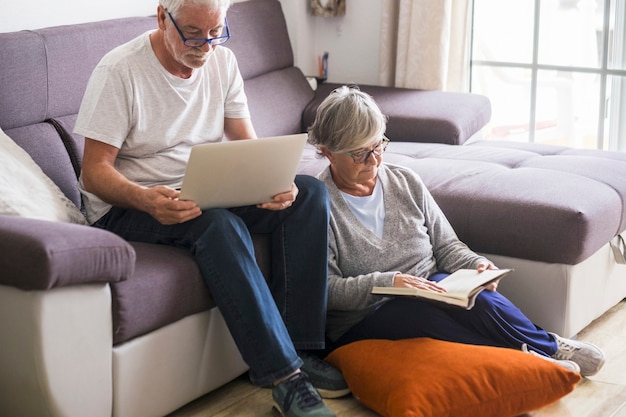 Para seniorów zrelaksowana i ciesząca się w domu na kanapie - dojrzała i emerytowana kobieta czytająca książkę w ciszy na ziemi i mężczyzna pracujący z laptopem lub komputerem PC