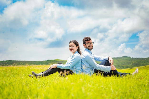 Para ślubna siedząca plecami do siebie na trawie Para ślubna w polu siedząca plecami do siebie patrząca w kierunku kamery