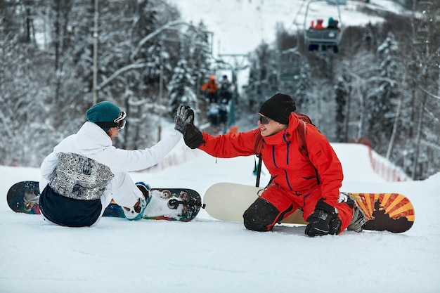Para snowboardzistów przybija sobie piątkę, siedząc na ośnieżonych szczytach stoku na tle błękitnego nieba i wyciągu narciarskiego w tle w zimowym ośrodku narciarskim w słoneczny dzień.