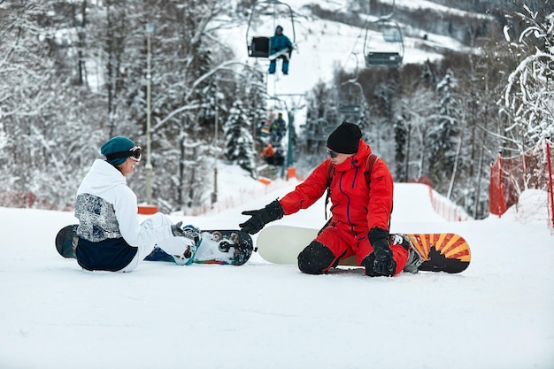 Para Snowboardzistów Przybija Sobie Piątkę, Siedząc Na Ośnieżonych Szczytach Stoku Na Tle Błękitnego Nieba I Wyciągu Narciarskiego W Tle W Zimowym Ośrodku Narciarskim W Słoneczny Dzień.