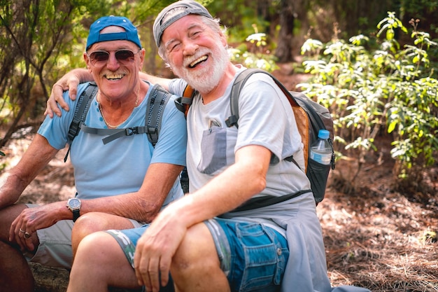 Para szczęśliwych starszych mężczyzn w górskiej wycieczce, obejmując patrząc na kamerę, śmiejąc się, siedząc, by odpocząć, ciesząc się zdrowym stylem życia w przyrodzie