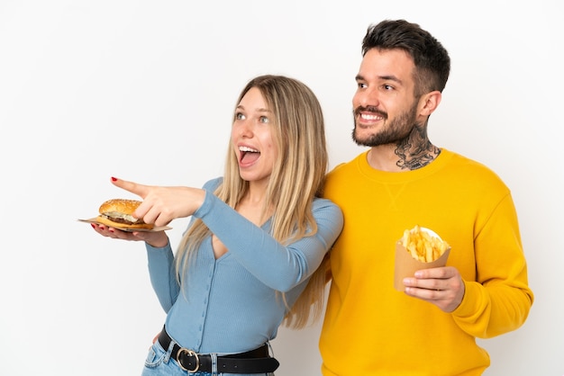 Para Trzymająca Hamburgera I Smażone Frytki Na Białym Tle, Wskazując Na Bok, Aby Zaprezentować Produkt