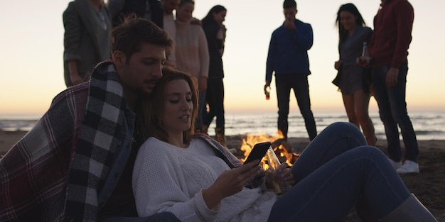 Para używająca telefonu komórkowego podczas imprezy na plaży z przyjaciółmi pijącymi piwo i dobrze się bawiącą