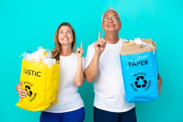 Para w średnim wieku trzymająca worki do recyklingu pełne papieru i plastiku na białym tle pokazujące i unoszące palec na znak najlepszych