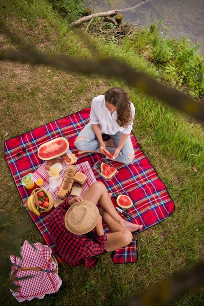 Para zakochanych, ciesząc się piciem i jedzeniem w czasie pikniku w pięknej przyrodzie na widok z góry na brzeg rzeki
