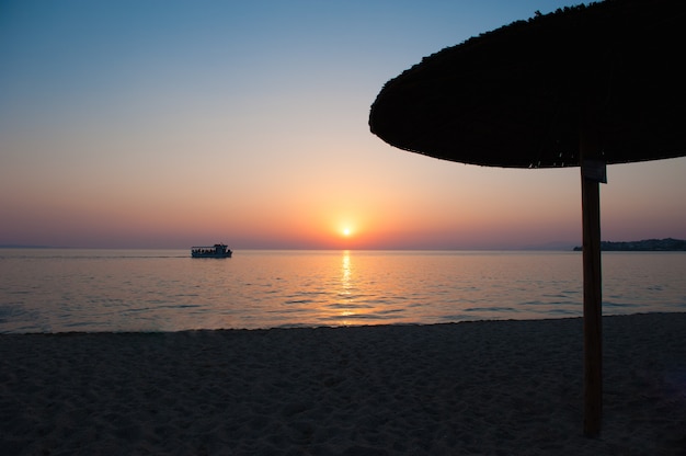 Parasole plażowe o zachodzie słońca, z leżakami, gorący zachód słońca. Miękkie fale morskie i pęcherzyki na plaży z zachodem słońca niebo. Żaglówka o zachodzie słońca w pobliżu plaży, parasol plażowy