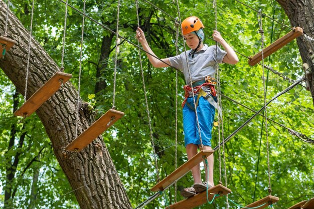 Park linowy Nastolatek chłopiec w kasku chodzi po podwieszonych drabinkach linowych Karabińczyki i pasy bezpieczeństwa Bezpieczeństwo Letnia aktywność Sport Plac zabaw dla dzieci w przyrodzie w lesie