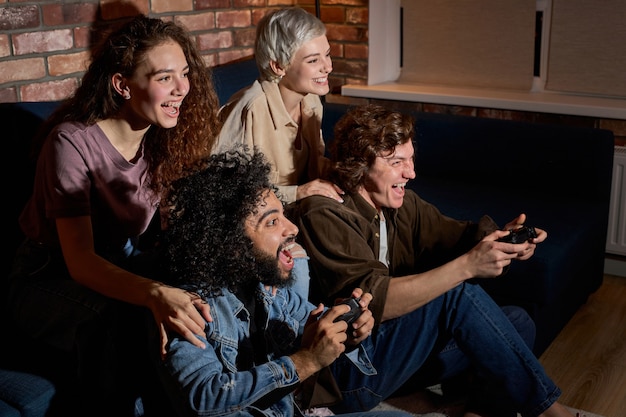 Pary przyjaciół trzymających joystick, grając w gry wideo na konsoli, aby się dobrze bawić, siedząc zrelaksowany. Szczęśliwi faceci i panie w strojach codziennych spędzają czas razem