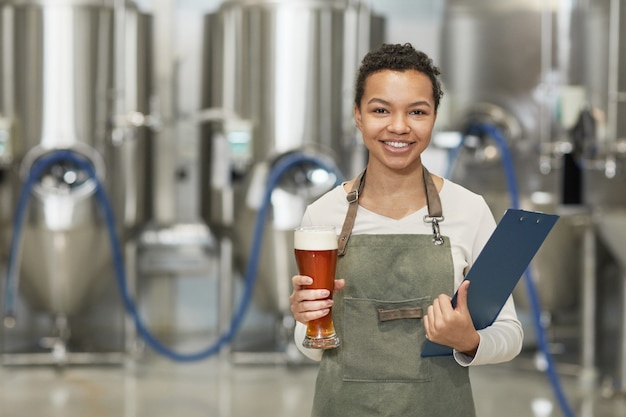 Pas w górę portret uśmiechniętej afroamerykańskiej kobiety trzymającej szklankę do piwa i patrząc na kamerę stojąc w warsztacie w fabryce browarnictwa, kopia przestrzeń