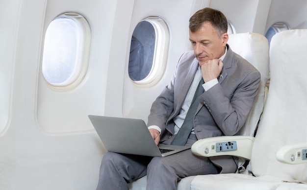 Pasażer kaukaski biznesmen w samolocie pracy z obszarem komputera przy użyciu połączenia bezprzewodowego na pokładzie, biznesmen kaukaski sprawdzanie pracy w kabinie samolotu.