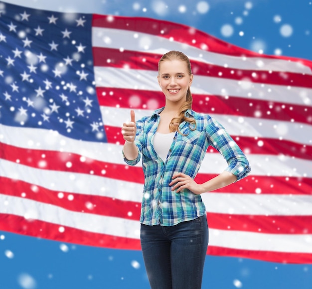 patriotyzm, edukacja, gest i koncepcja ludzi - uśmiechnięta dziewczyna w zwykłych ubraniach pokazująca kciuk w górę nad amerykańską flagą i niebieskim niebem na tle śniegu