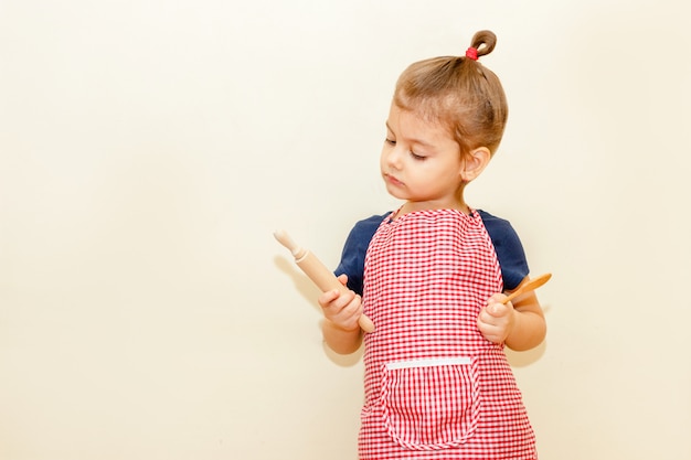 Patrzeć z zainteresowaniem małej dziewczynki z szefa kuchni fartuchem trzyma drewnianą toczną szpilkę i łyżkę na beżowym tle