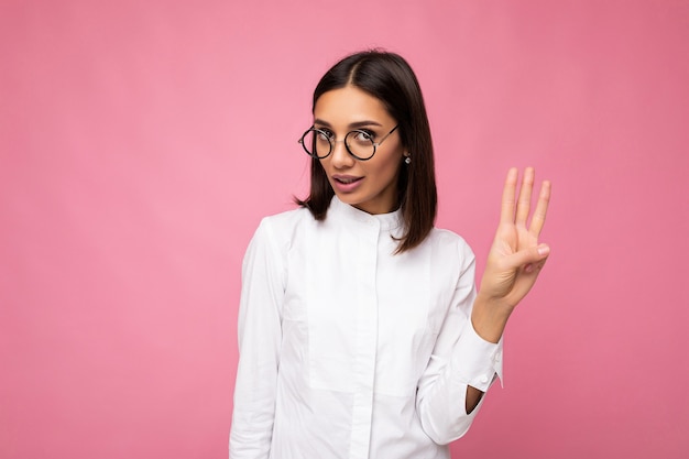 Pchnięcie fotografii pięknej młodej kobiety brunetka ubrana w ubranie i stylowe okulary optyczne na białym tle nad kolorowym patrząc na kamery i pokazując trzy palce.