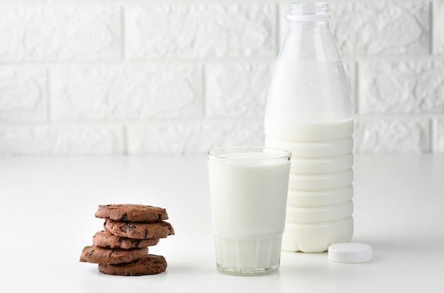 Pełen szklany kubek mleka i przezroczysta plastikowa butelka z mlekiem, obok stosu okrągłych ciasteczek z kawałkami czekolady na białym stole