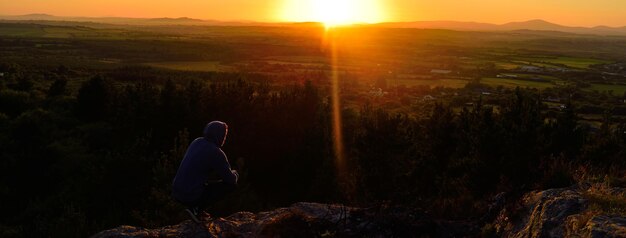 Zdjęcie pełna długość człowieka przykucniętego na klifie na tle krajobrazu podczas zachodu słońca