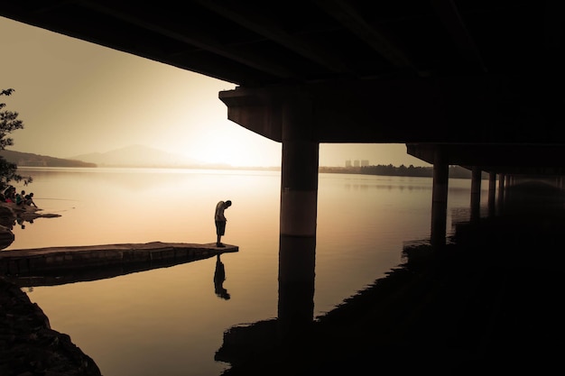 Pełna długość człowieka stojącego na krawędzi molo w jeziorze podczas zachodu słońca