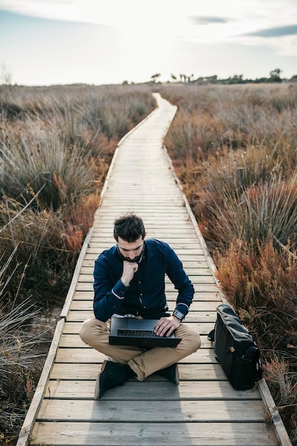 Zdjęcie pełna długość człowieka używającego laptopa podczas siedzenia pośród pola na promenadzie