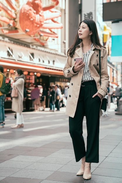 pełna długość japońskiej bizneswoman korzystającej ze smartfona z aplikacją mapy online wyszukującej sposoby odwiedzenia słynnej lokalnej restauracji. Szczęśliwy asian kobieta pracownik stojący na ulicy dotonbori i znaleźć obiad.
