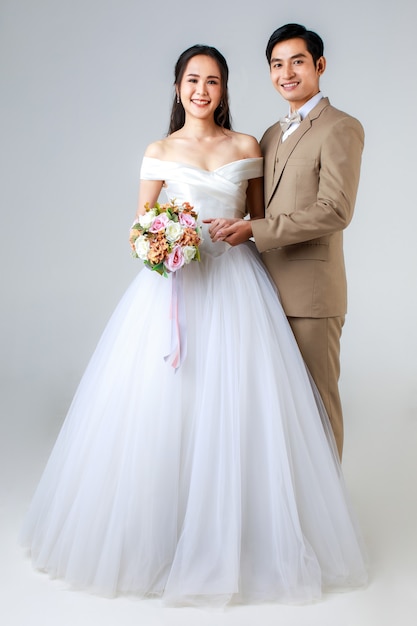 Pełna długość młoda atrakcyjna para azjatyckich, mężczyzna ubrany w beżowy garnitur, kobieta ubrana w białą suknię ślubną stojącą razem trzymając się za ręce. Koncepcja fotografii przedślubnej.