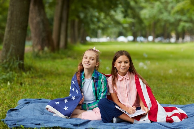 Pełna długość Portret dwóch ślicznych dziewczyn objętych amerykańską flagą siedzi na kocu piknikowym w parku i uśmiecha się