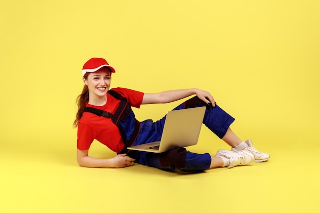 Pełna długość portret poręcznej kobiety leżącej na podłodze ze szczęśliwym wyrazem twarzy, trzymając laptopa do obsługi klienta online, centrum kontaktu z serwisem naprawczym. Kryty strzał studio na białym tle na żółtym tle