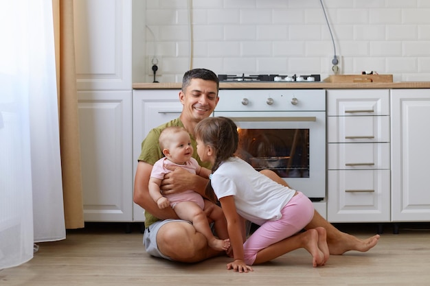 Pełna długość portret szczęśliwego śmiejącego się człowieka noszącego t-shirt w stylu casual, siedzącego na podłodze w kuchni z córkami słodkie dziewczyny bawiące się z ojcem, bawiące się śmiejąc się