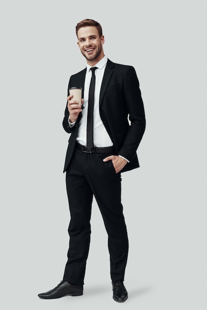 Pełna długość przystojnego młodego mężczyzny w stroju formalnym, patrzącego na kamerę i uśmiechającego się, stojąc na szarym tle