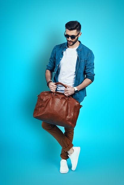 Pełna długość przystojnego stylowego młodego mężczyzny dostosowującego torbę stojącą przed niebieską ścianą
