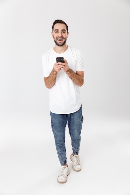 Pełna długość przystojnego, wesołego mężczyzny noszącego pustą koszulkę stojącego na białym tle nad białą ścianą, korzystającego z telefonu komórkowego
