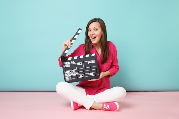 Pełna długość strzału kobieta w różowej koszuli białych spodniach siedząca na podłodze z klapką do robienia filmów