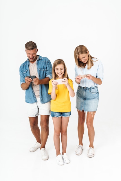 Pełna długość wesołej podekscytowanej rodziny noszącej swobodny strój stojący na białym tle, grający w gry na telefonach komórkowych