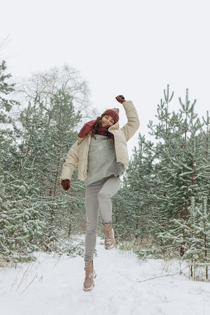 Pełna długość zachwyconej kobiety w ciepłej odzieży wierzchniej skaczącej w śnieżnym lesie i cieszącej się zimą