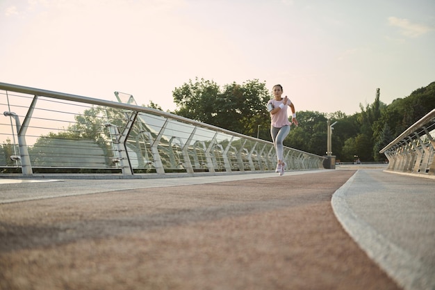 Pełna długość. Zdeterminowana kobieta jogger sportowiec biega na bieżni wzdłuż mostu, wykonuje poranny trening cardio o świcie. Trening sportowy na świeżym powietrzu, odchudzanie, sport, fitness, koncepcja aktywnego stylu życia