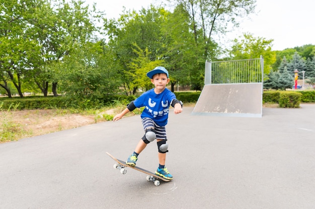 Pełna długość zdeterminowanego chłopca noszącego ochraniacze i czapkę wykonującą proste sztuczki na deskorolce w skateparku w letni dzień