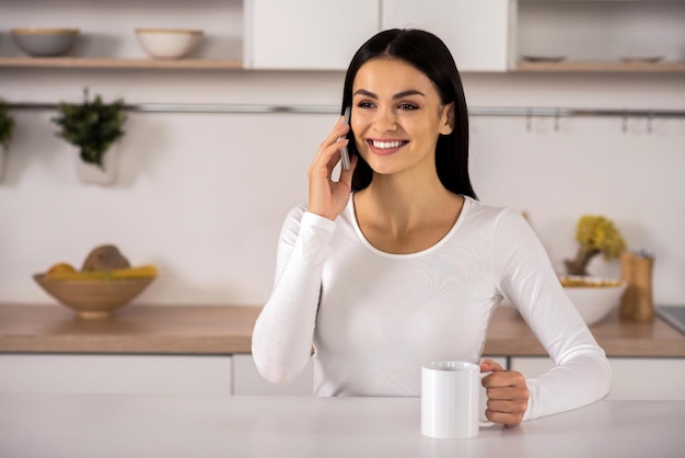 Pełna radości Wesoła, atrakcyjna młoda kobieta rozmawia przez smartfon siedząc w kuchni