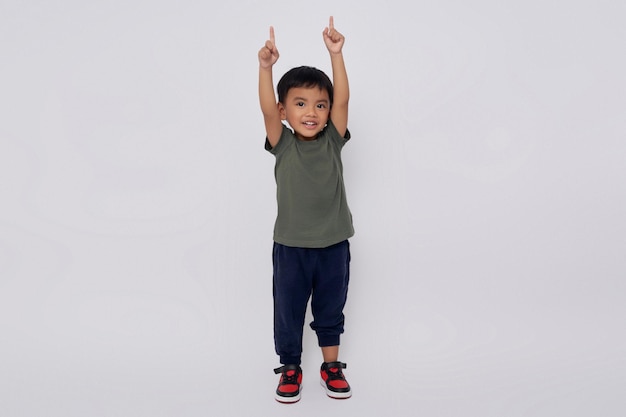 Zdjęcie pełne ciało mały uśmiechnięty szczęśliwy azjatycki chłopiec 23 lat noszący zieloną koszulkę stojący pewnie podczas wykonywania gestu zwycięzcy odizolowany na białym tle
