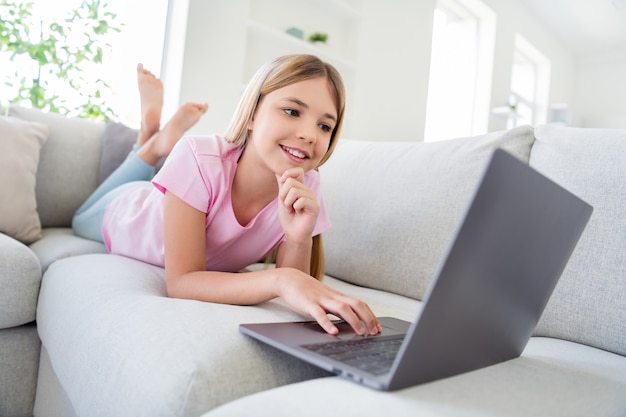 Pełne zdjęcie pozytywnej dziewczyny dziecko studiuje zdalne użytkowanie laptopa ma projekt online na czacie, komunikację z kolegami z klasy, leżącą kanapą w domu w pomieszczeniu