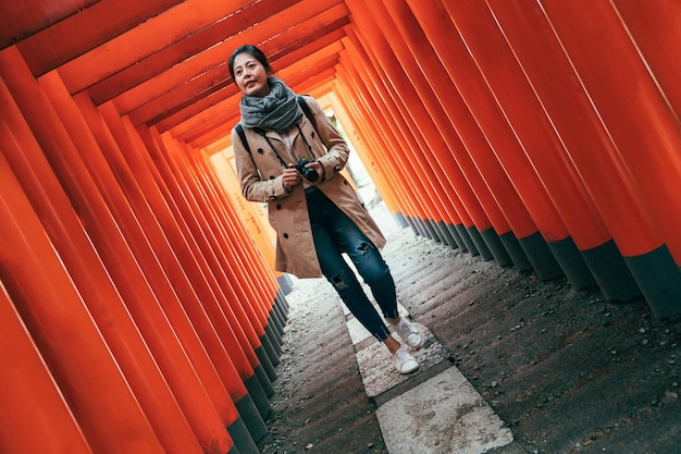 pełnej długości azjatycki fotograf radośnie idący przez czerwone bramy torii w świątyni tenmangu w japońskiej osace. młoda dziewczyna odwiedzając japońskie shinto w wiosennych podróżach wakacyjnych. piękna kobieta z aparatem